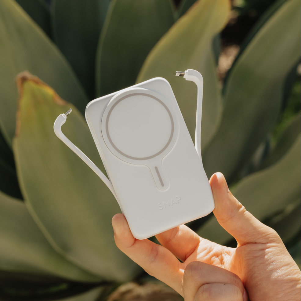 Hand holding a white electronic device with a cable against a green plant background